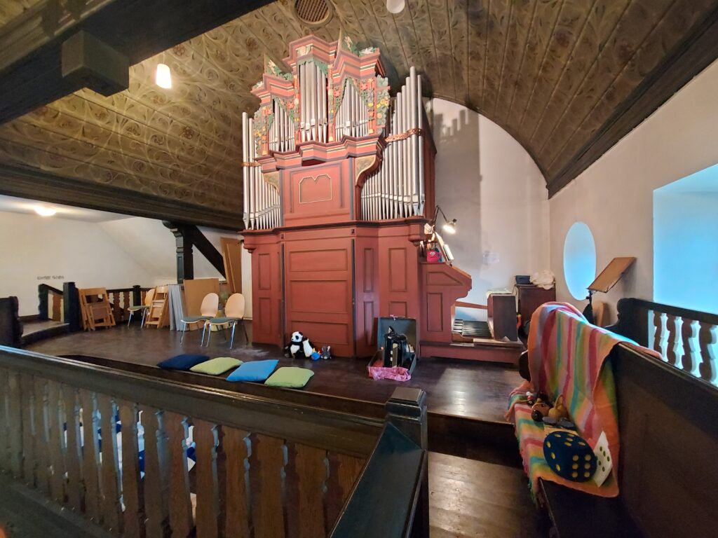 Blick auf eine Orgel, davor steht ein Akkordeon und Kuscheltiere sitzen am Boden. Es sind bunte Kissen zu sehen, und auf einer Kirchenbank liegt ein großer Würfel, daneben Musikinstrumente. Am Spieltisch der Orgel eine rote Kaffeetasse aus Plüsch.