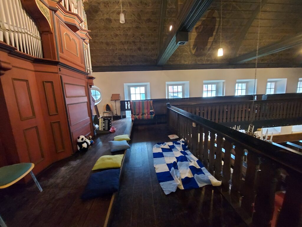 Wir befinden uns auf einer Orgelempore. Links im Bild die Orgel, davor sitzen Kuscheltiere und ein Akkordeon ist zu sehen. Es liegen Kissen auf den Stufen und eine bunte Kuscheldecke. 